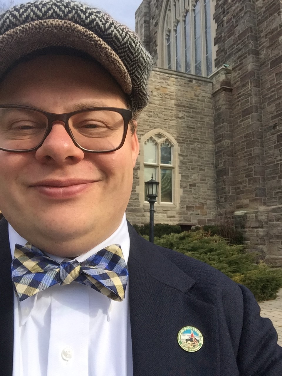 A close up of Tay Moss, a smiling man in glasses, a newsboy cap and checkered bowtie. Behind him is a church building.