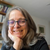 A headshot of Helen Blier, a white woman with long gray hair and glasses. She is resting her chin on her palm and smiling.